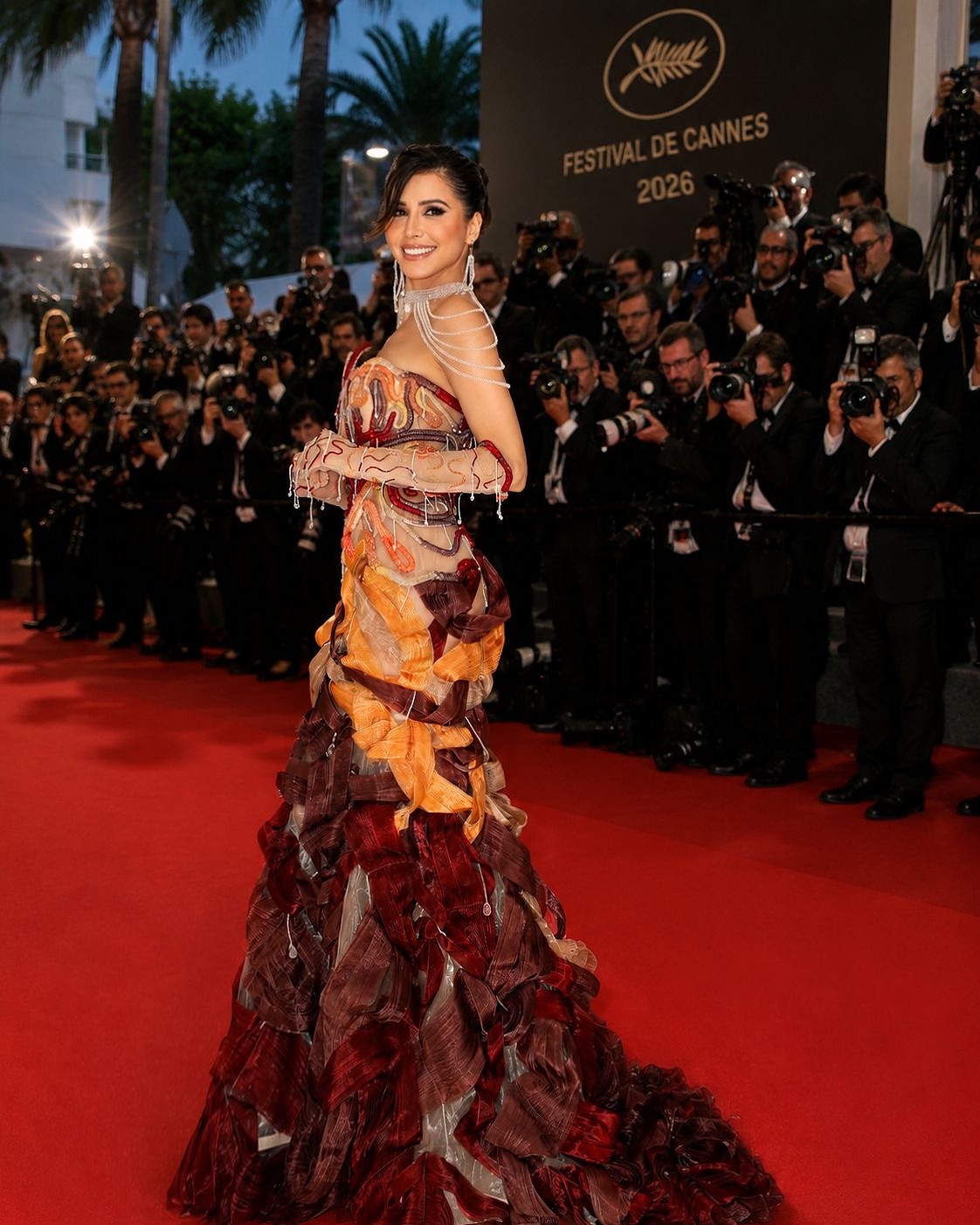 Somayeh sur le tapis rouge du Festival de Cannes 2026, en robe sculptée bordeaux et orange, devant les photographes