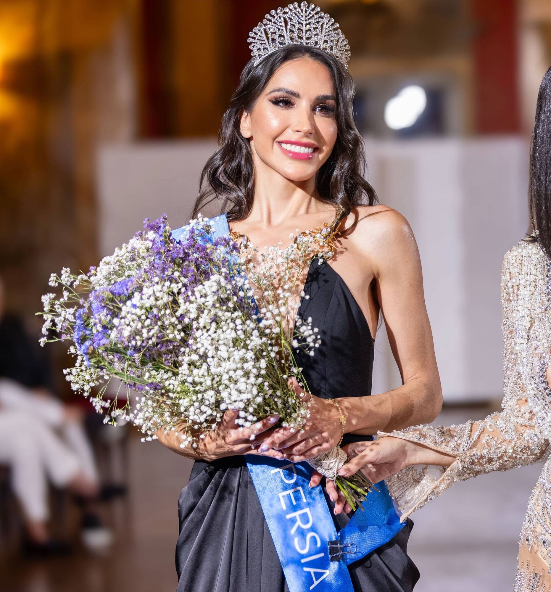 Somayeh couronnée, en robe noire, écharpe bleue Miss Universe Persia, un bouquet de gypsophile et de lavande à la main